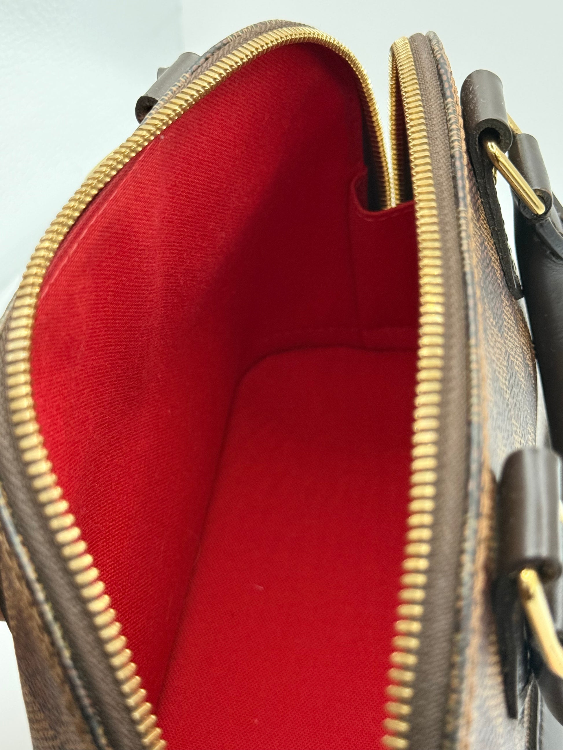 Close-up of an open Louis Vuitton Alma BB handbag showing the interior red lining, brown monogram canvas exterior, and gold-tone zippers and hardware.