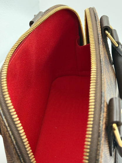 Close-up of an open Louis Vuitton Alma BB handbag showing the interior red lining, brown monogram canvas exterior, and gold-tone zippers and hardware.