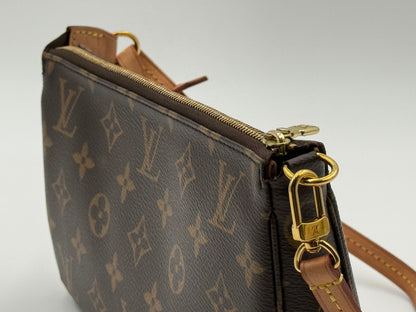 A close-up of a Louis Vuitton monogram canvas wristlet pouch with brown leather trim and gold-tone hardware, set against a neutral light gray background. Pochette Accesoires.