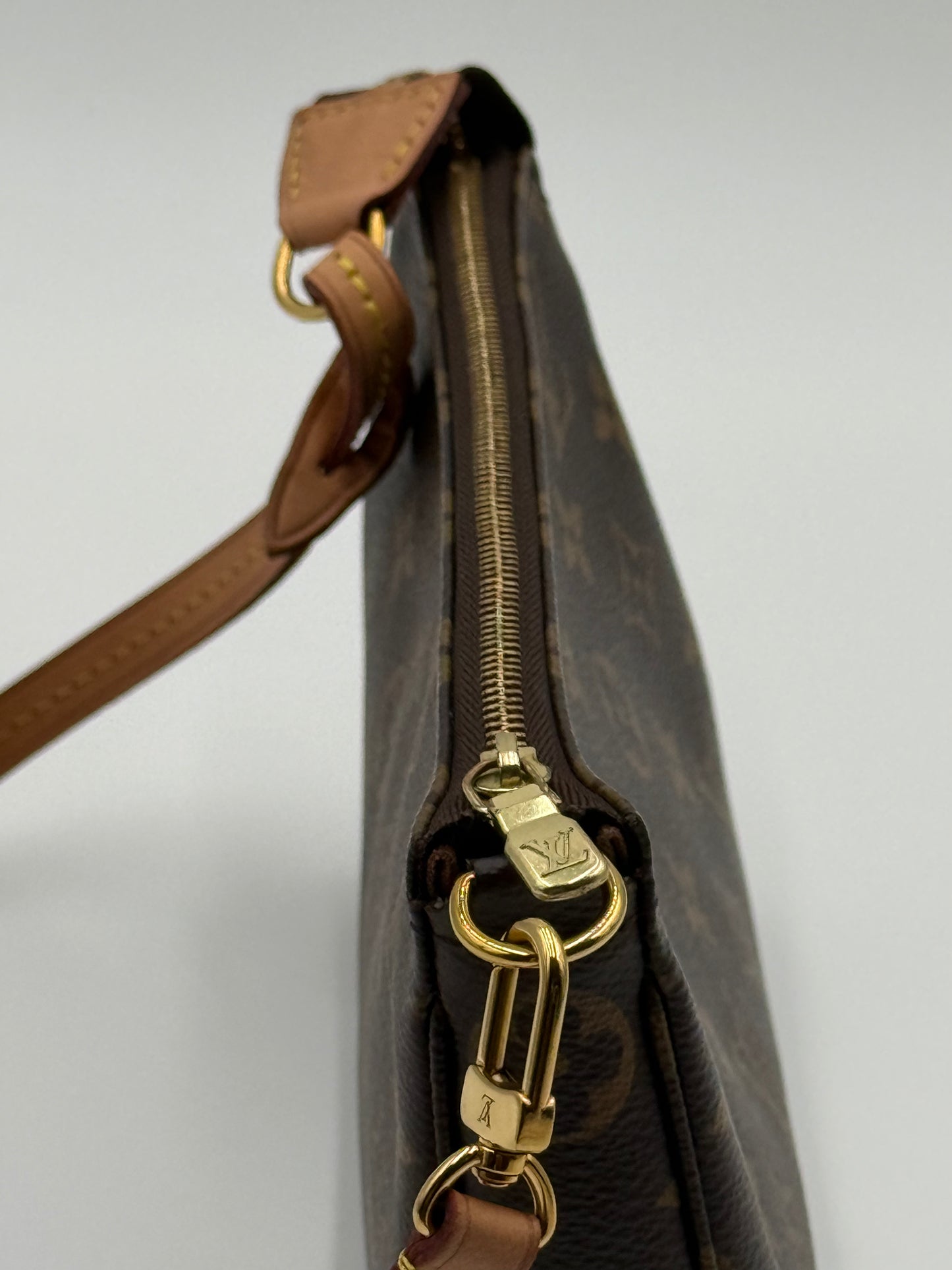 Close-up of a brown monogram handbag with gold-tone hardware and a tan leather strap on a light gray background. Pochette Accessoires.
